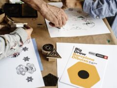 Se puede apreciar una mesa de madera llena de papeles en blanco. En las hojas en blanco hay dibujos y estampas de distintas formas. Tamvién hay manos de niños y manos de adultos, que son las personas que están realizando el taller. Es un plano detalle.