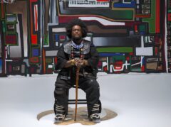 El saxofonista y compositor estadounidense Kamasi Washington está sentado en una silla. Se encuentra en medio de una sala vacía con papel pintado. EL papel pintado se asemeja a los colores y a los patrones de su camiseta.