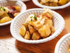 Se aprecia algunos platos de papel llenos de una famosa tapa española. Son patatas bravas con salsa y condimentos. Al fondo, se distingue una mesa rústica de madera.