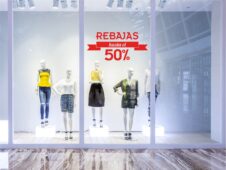 Escaparate con maniquíes vestidos con gamas azuladas y amarillas. El escaparate es blanco y los maniquíes también. Tienen una silueta femenina. En el cristal se aprecia la palabra 'Rebajas'.