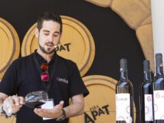 Un profesional agrario prueba vino. Tiene un micrófono de diadema y tras él, vemos diferentes barriles de vino en los que se ve el logo de 'Fira àpat'.